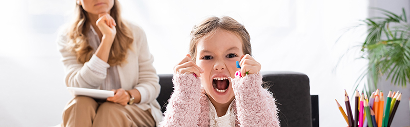 little girl patient screaming and clenching figures while visiti Child with autism expressing frustration during an ABA therapy session, with a therapist in the background. Supportive ABA therapy in Los Angeles County to help children manage challenging behaviors.